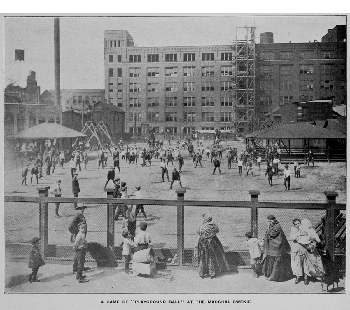Children play baseball inside a playground with large buildings in the background