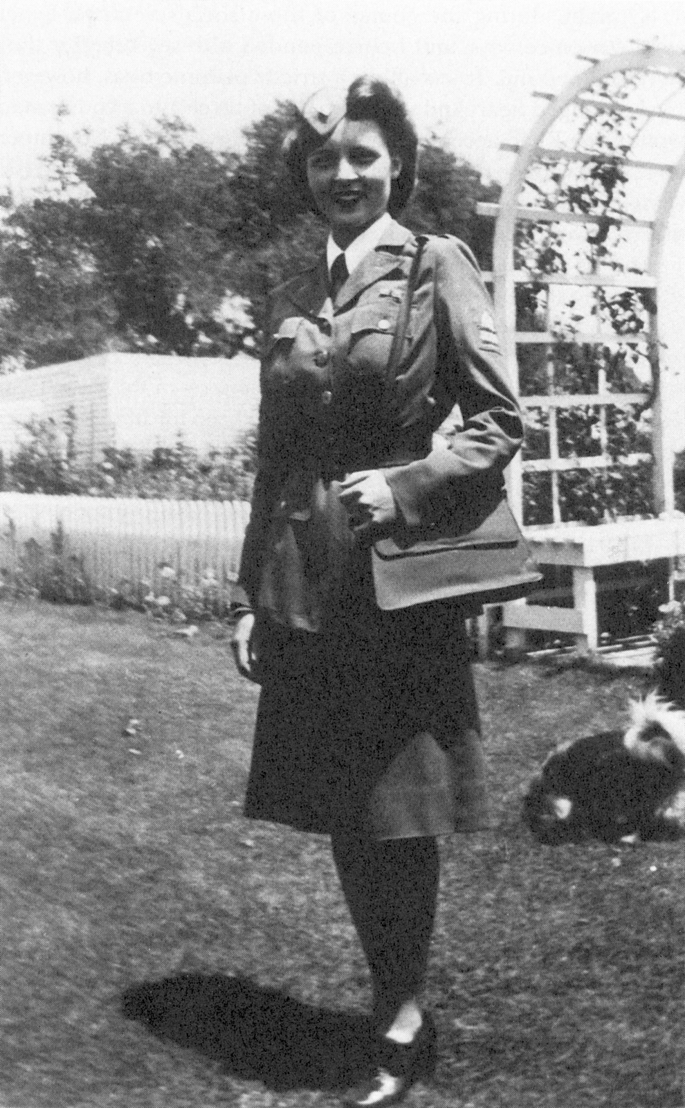 Black-and-white photograph of a young Betty White wearing her American Women’s Voluntary Services uniform, complete with shoulder bag, while standing outdoors.