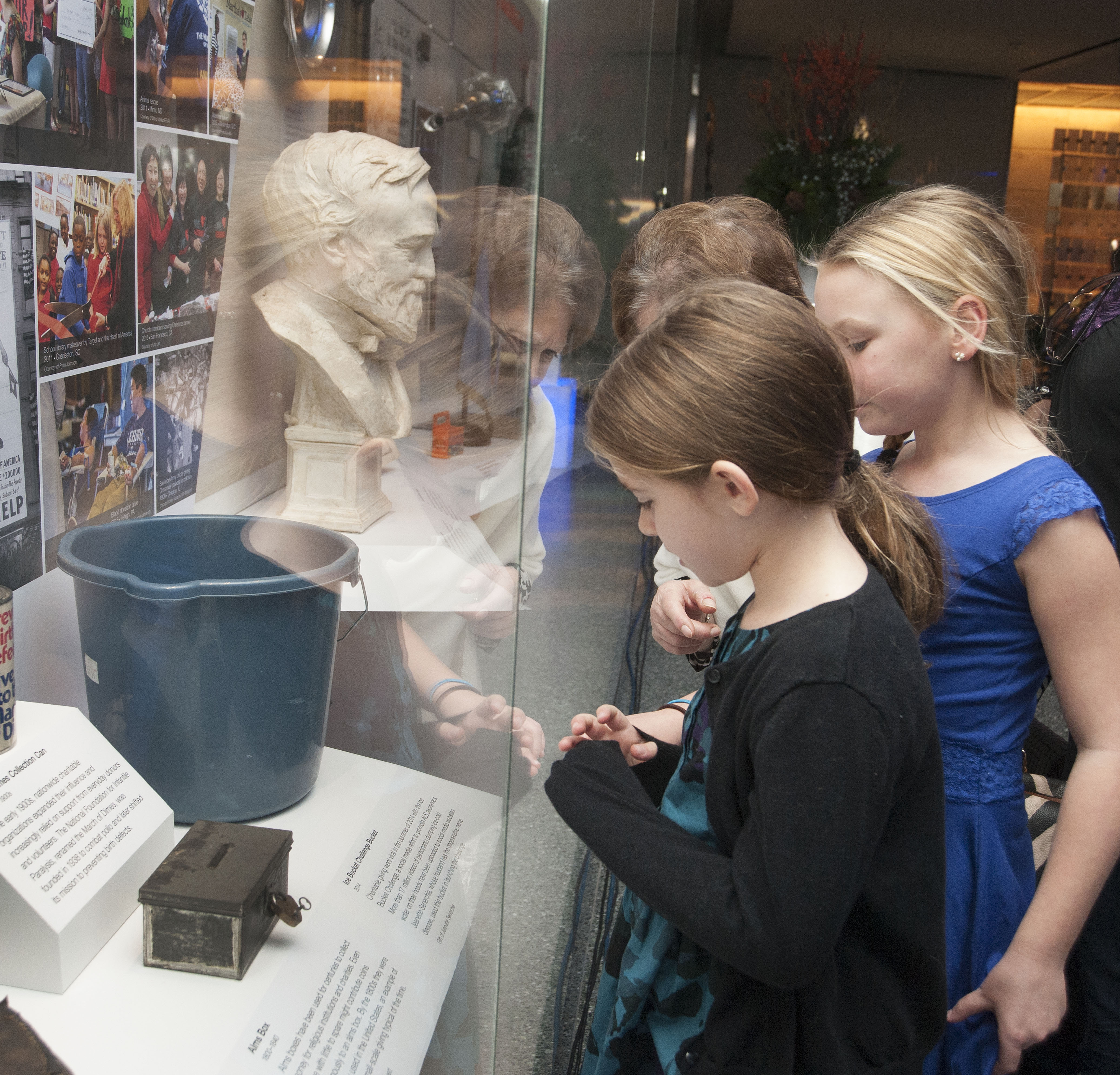Two young girls look through the glass of a museum display to gaze at a blue bucket among other objects like a pair of boots or a bust of a man.