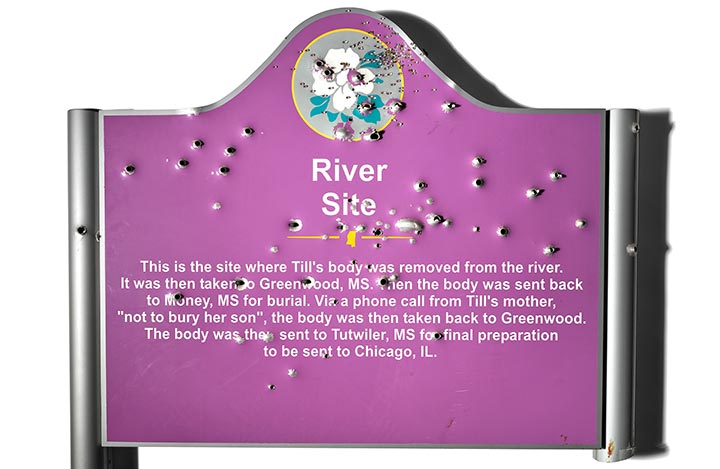 A metal historical marker professionally photographed against a white background. The sign’s purple paint and white lettering are disfigured and pierced by hundreds of pellet gun and bullet holes.