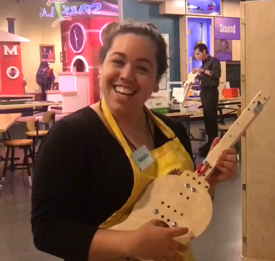 A photograph of a smiling woman in a yellow apron holding a makeshift wooden banjo