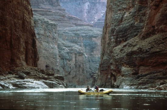 Photo of two people in a small yellow raft, wearing life preservers. They are on the water among very high-walled rock cannons.