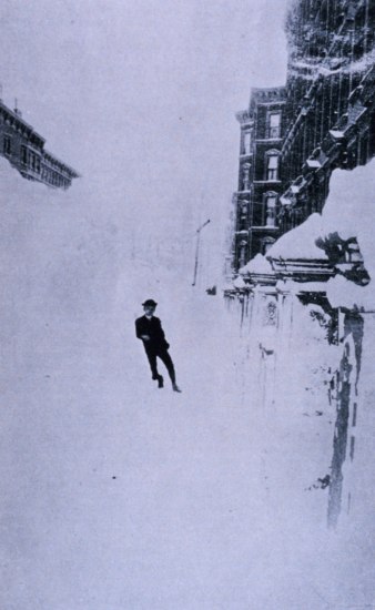 Black and white photo of man wearing a hat leaning against a giant bank of snow. To the right, snow-covered gate or fence and three story building. 