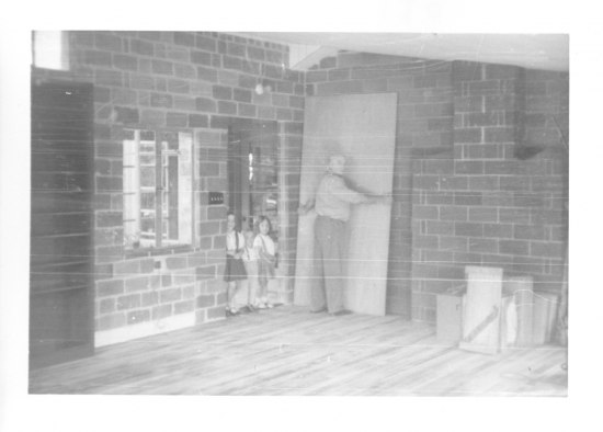 A black and white photograph of a man facing a wall and holding a large sheet of wood in a room with wood floors and brick walls. Two children stand in the doorway next to him.