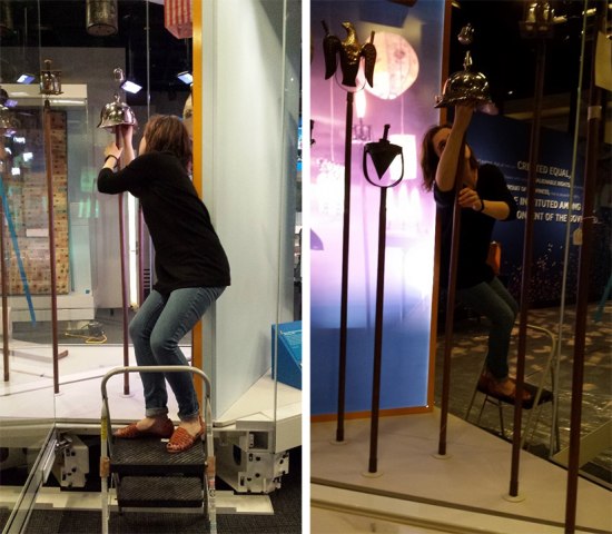 Two images. In both, a museum staff member stands on a step ladder, leaning to the side in order to adjust the placement of a campaign torch topper on a pole.