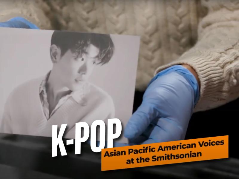 Gloved museum staff holding a photo of K-Pop artist Eric Nam, accompanied by the text "K-Pop: Asian Pacific American Voices at the Smithsonian"